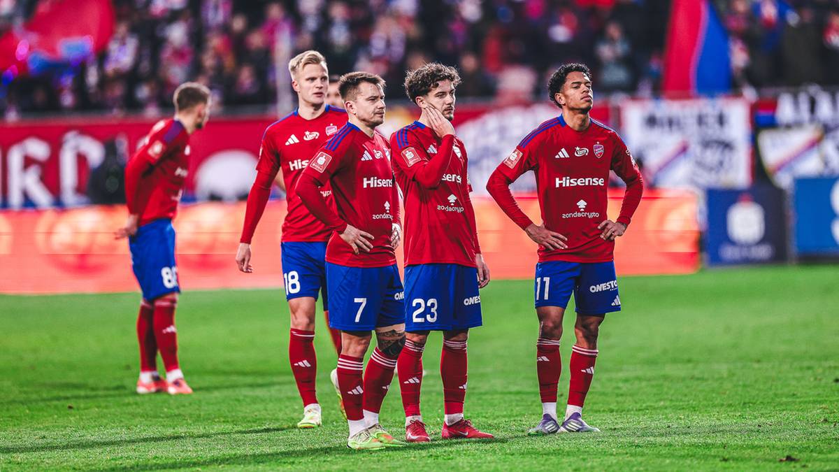 Pięciu piłkarzy w czerwonych strojach z niebieskimi detalami stoi na zielonej murawie stadionu piłkarskiego, patrząc w r&oacute;żnych kierunkach.