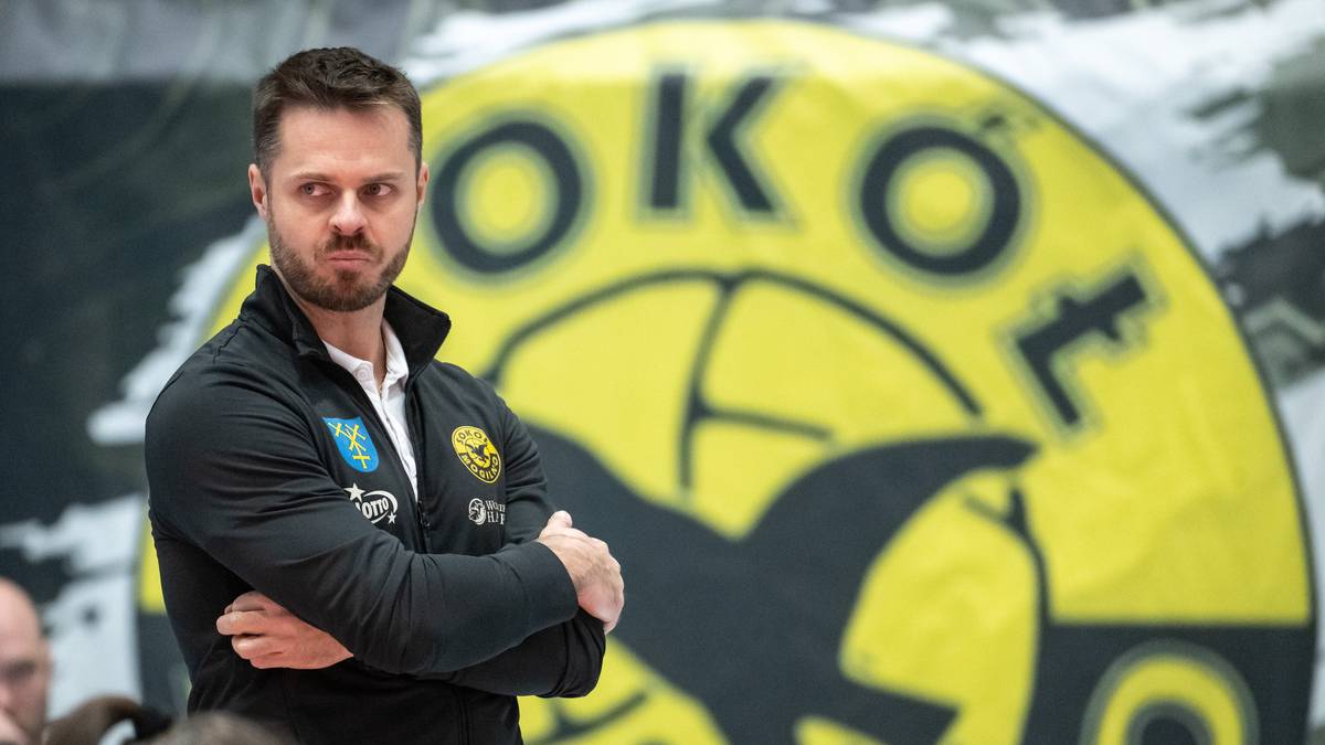 Mężczyzna w czarnej kurtce sportowej stoi ze skrzyżowanymi rękami przed ż&oacute;łtym emblematem klubu sportowego.