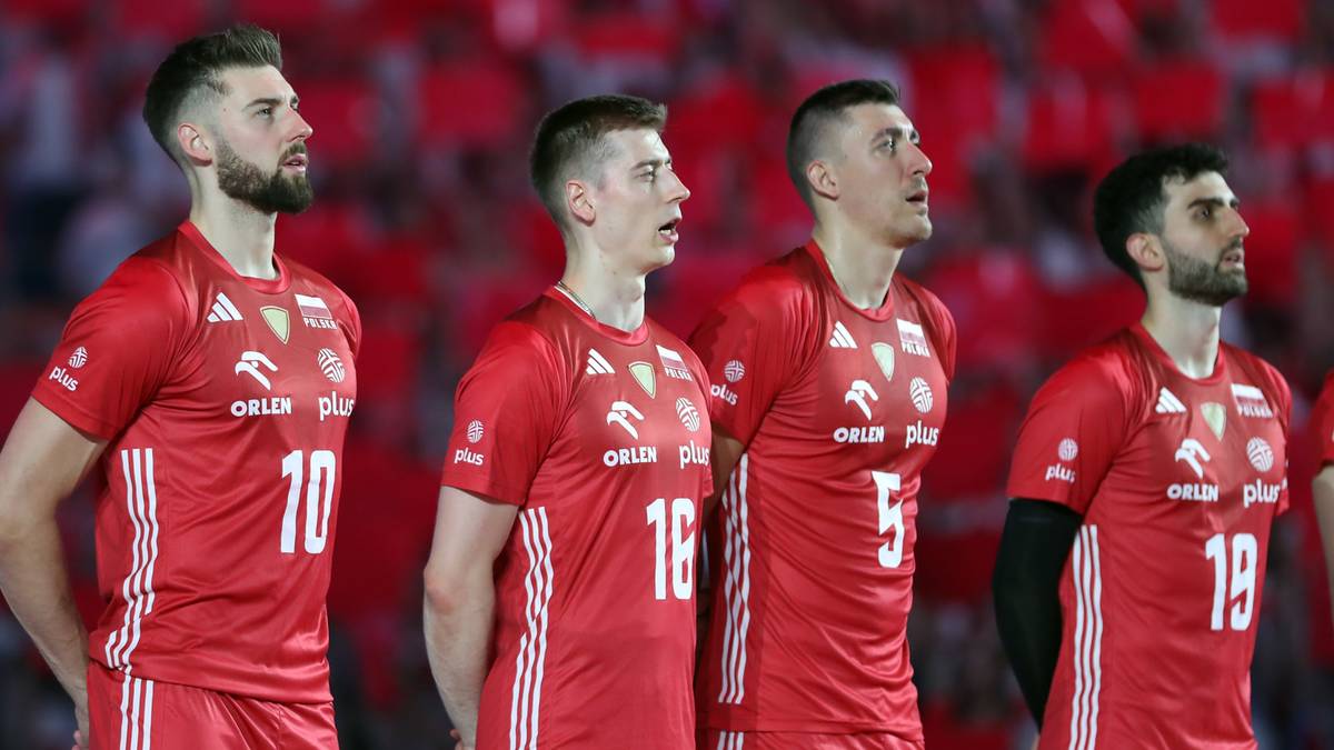 Czterech siatkarzy w czerwonych koszulkach sportowych z białymi numerami na plecach, stojących obok siebie na tle trybun wypełnionych kibicami.