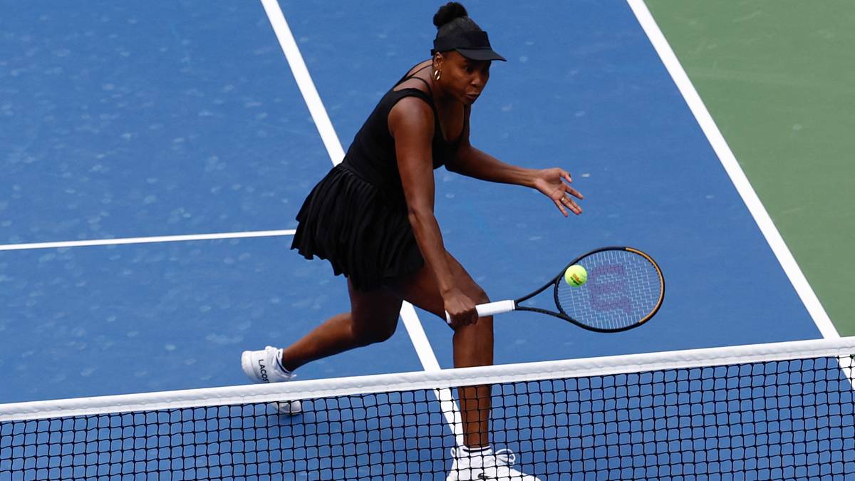Tenisistka Venus Williams w czarnej sukience i czapce z daszkiem uderza rakietą piłkę do tenisa na niebieskim korcie.