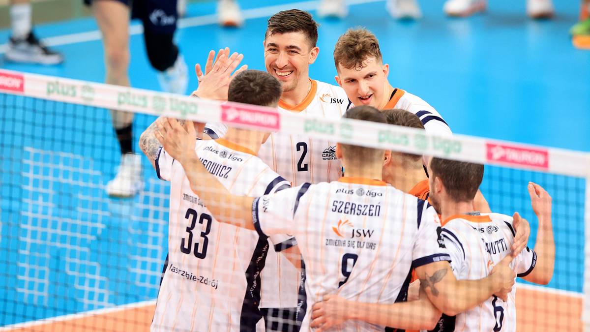 Siatkarze w białych strojach z pomarańczowymi detalami cieszą się po meczu, przytulając się do siebie na boisku.