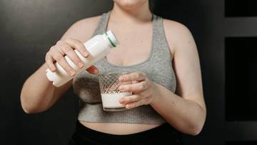 Kobieta w szarym topie sportowym nalewa biały płyn z butelki do szklanki.