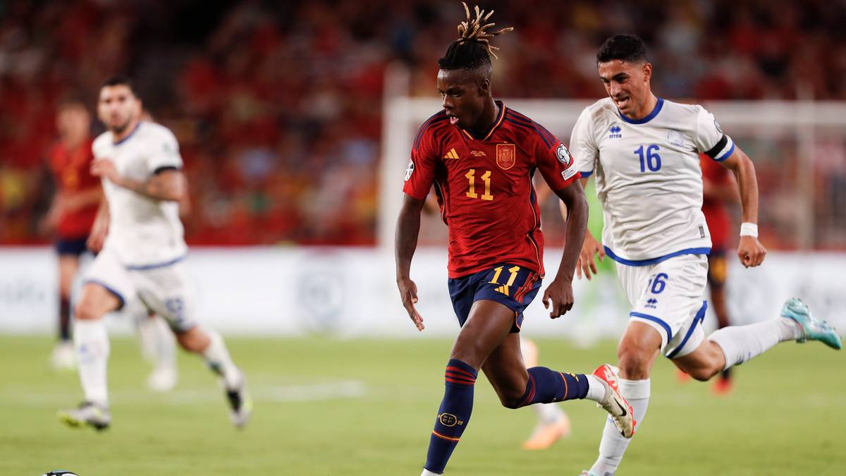 Piłkarz w czerwonej koszulce z numerem 11 na plecach biegnie z piłką po boisku, obok niego biegnie piłkarz w białej koszulce z numerem 6. W tle inni zawodnicy i kibice na trybunach.