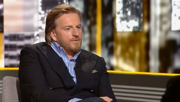 Mężczyzna z jasnymi włosami i brodą, ubrany w niebieską koszulę w paski i marynarkę, siedzi w fotelu.