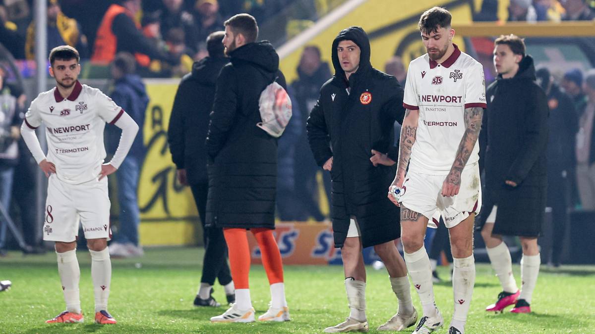 Piłkarze w białych strojach stoją na boisku, w tle widać kibic&oacute;w na trybunach stadionu.