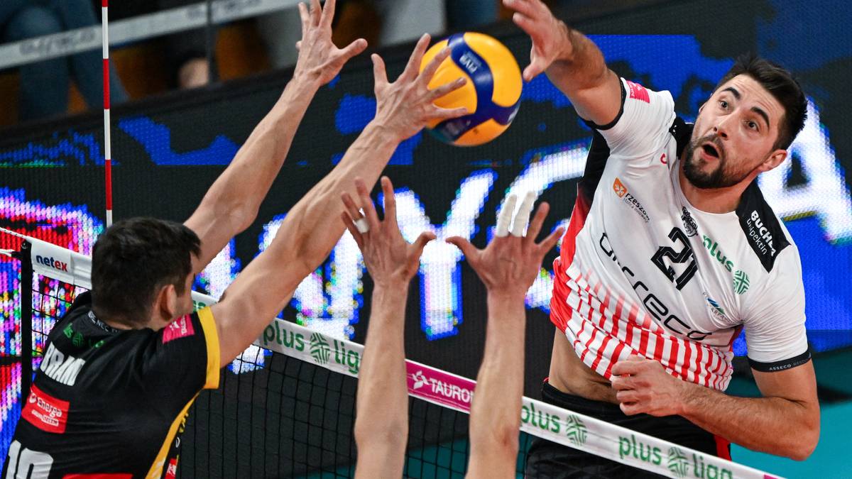 Siatkarz w białej koszulce z numerem 21 w białej i czerwonej koszulce próbuje wykonać atak, podczas gdy dwaj gracze w czarnych strojach próbują go zablokować. Piłka do siatkówki jest widoczna w powietrzu.