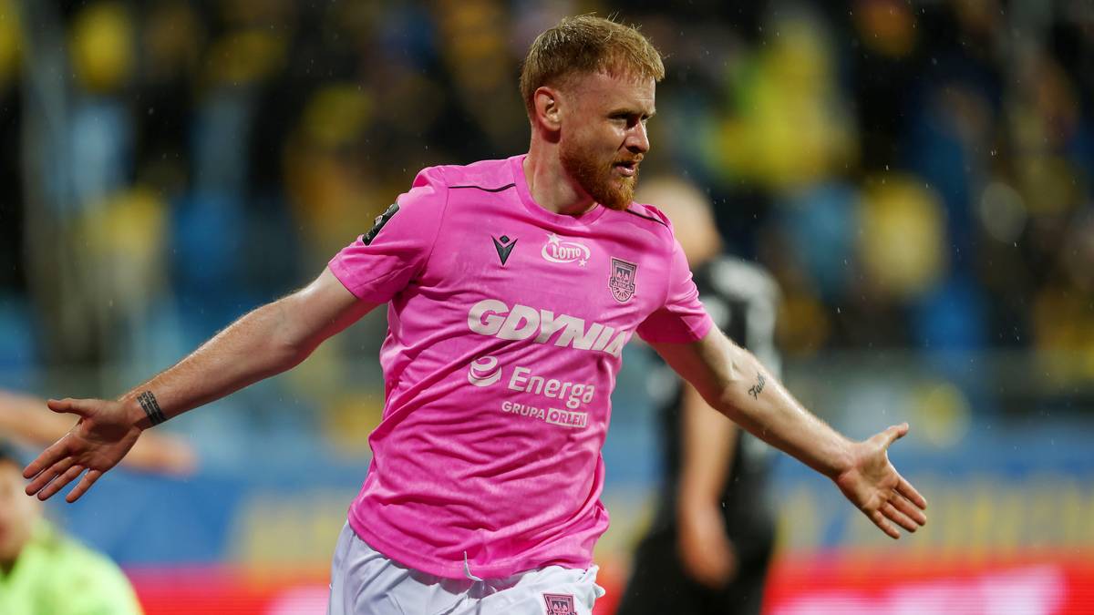 Piłkarz w r&oacute;żowej koszulce Arki Gdynia, z uniesionymi ramionami, świętujący bramkę na stadionie.