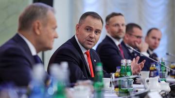 Mężczyzna w garniturze i czerwonym krawacie przemawia przy stole konferencyjnym, wskazując na problem.