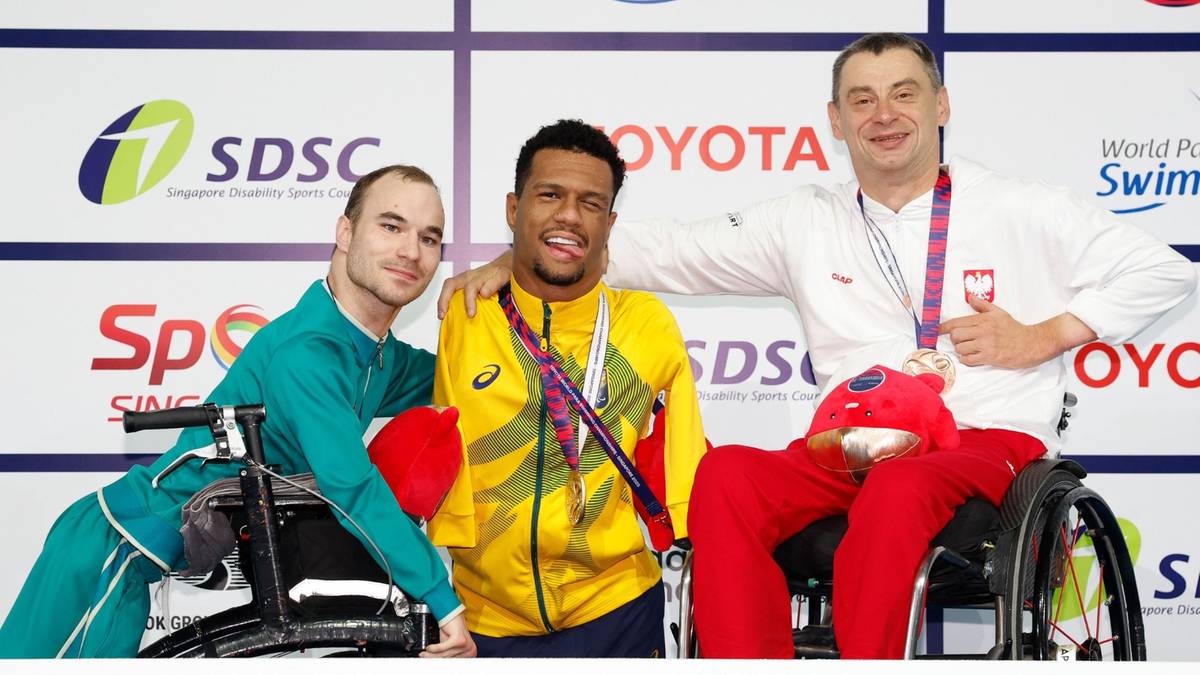 Trzech mężczyzn pozuje do zdjęcia na podium. Dwóch z nich siedzi na wózkach inwalidzkich. Jedna osoba ma medal na szyi.