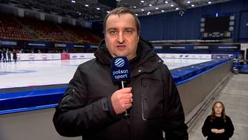 Mężczyzna w czarnej kurtce stoi na lodowisku i trzyma mikrofon z logo Polsat Sport. W tle widać trybuny lodowiska.