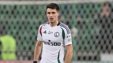 Piłkarz Legii Warszawa w białej koszulce, czarnych spodenkach i pomarańczowych butach stoi na boisku piłkarskim.