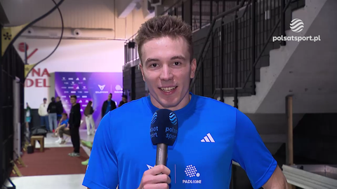 Młody mężczyzna Jakub Szymański, ubrany w niebieską koszulkę z logo Polsat Sport, uśmiecha się i m&oacute;wi do mikrofonu. W tle widać innych ludzi i fragmenty hali sportowej.