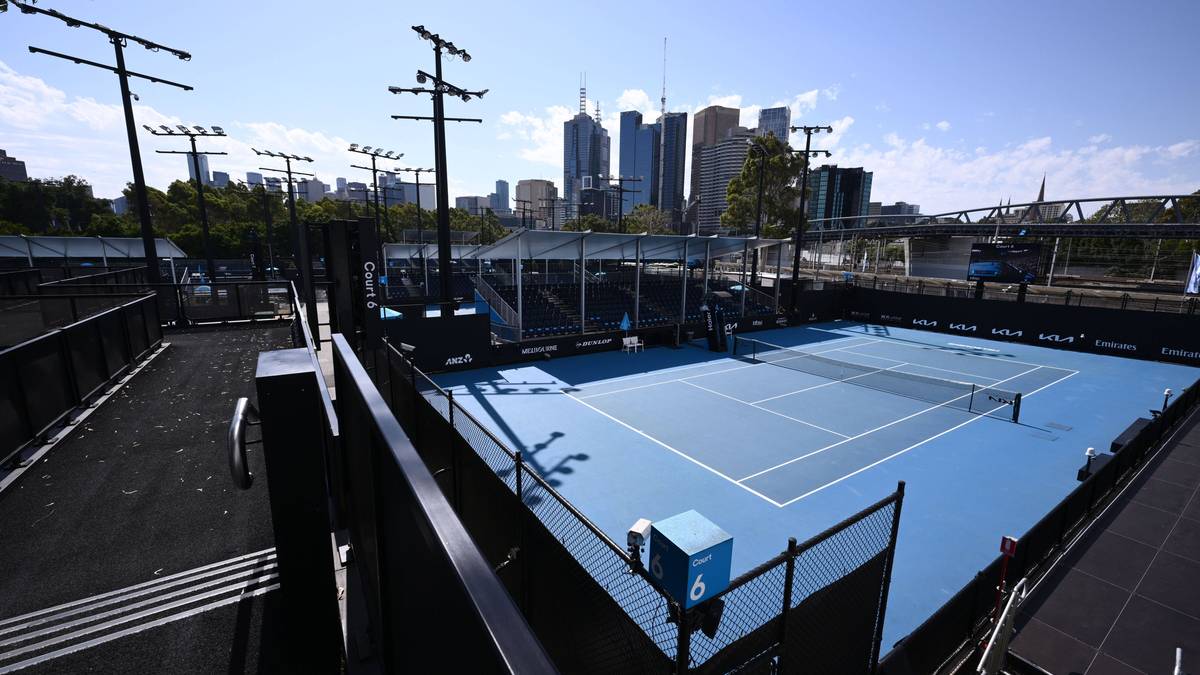 Widok na kort tenisowy z niebieską nawierzchnią i siatką, otoczony ogrodzeniem i słupami oświetleniowymi. W tle widać panoramę miasta z wieżowcami.