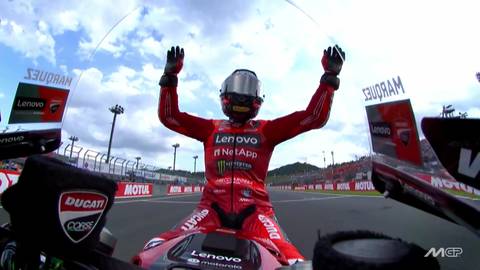 Motocyklista w czerwonym kombinezonie i kasku z uniesionymi rękami, na torze wyścigowym.