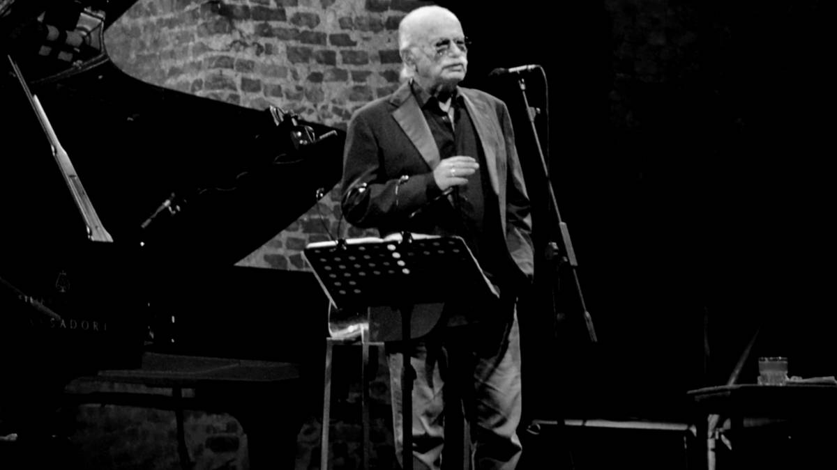 Starszy mężczyzna w okularach i garniturze stoi na scenie, trzymając dłoń przy twarzy, obok pulpitu z nutami i mikrofonu.