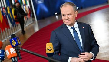 Donald Tusk stoi przed mikrofonami, w tle flagi państwowe.