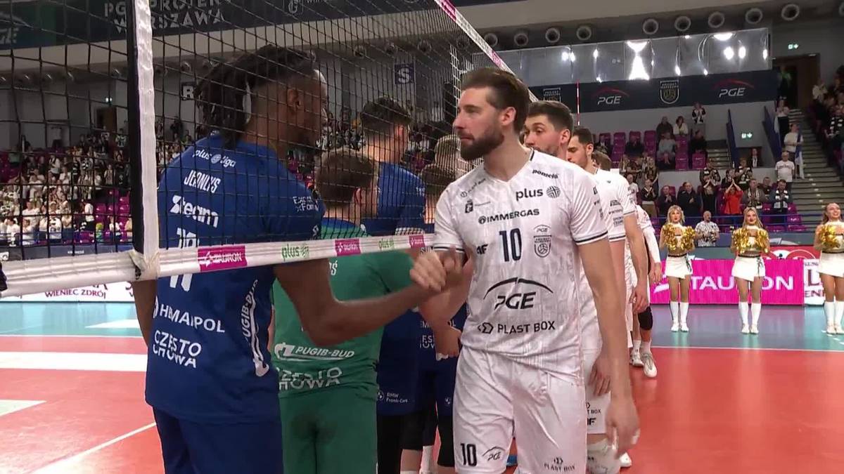 Dw&oacute;ch siatkarzy po meczu przy siatce, jeden w białej koszulce z numerem 10, drugi w granatowej, podaje sobie ręce. W tle kibice.