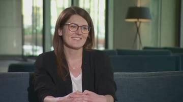 Kobieta w okularach i czarnej marynarce siedzi, uśmiechając się.