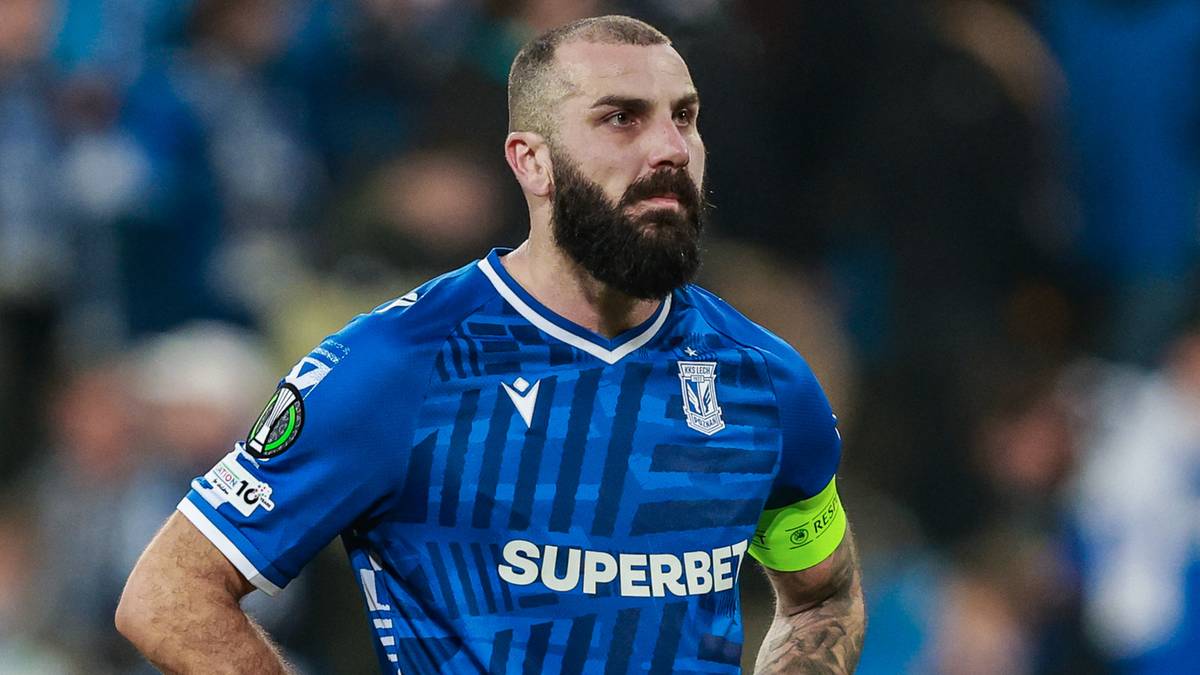 Mikael Ishak, kapitan Lecha Poznań, w niebieskiej koszulce z opaską na ramieniu, stoi na boisku piłkarskim.