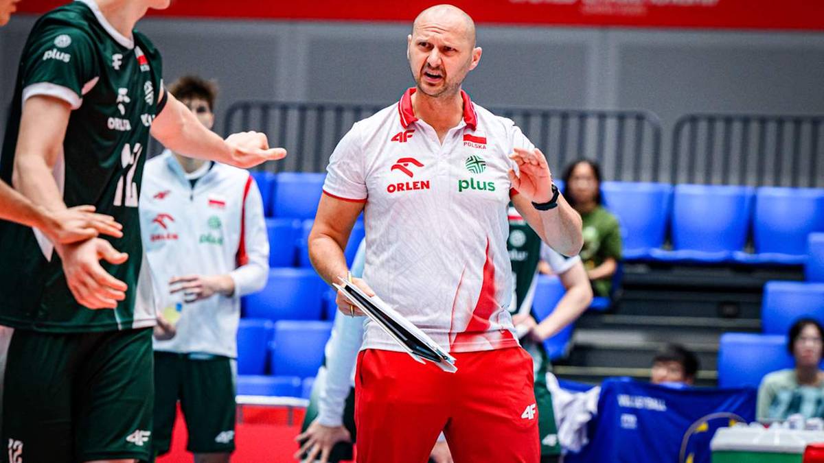 Trener w białej koszulce z czerwonymi elementami i napisem "PLUS" gestykuluje i przemawia do zawodników drużyny siatkówki.