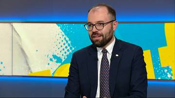 Mężczyzna w okularach i garniturze z krawatem, siedzący przed kolorowym tłem.