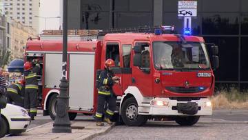 Samochód strażacki z włączonymi sygnałami świetlnymi, obok niego strażacy w akcji.