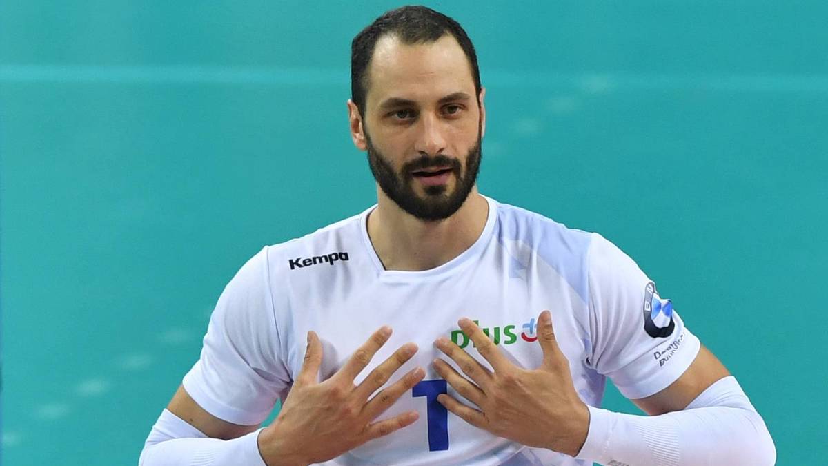 Mężczyzna z brodą i krótkimi włosami, ubrany w białą koszulkę sportową z długimi rękawami, patrzy przed siebie z poważnym wyrazem twarzy.