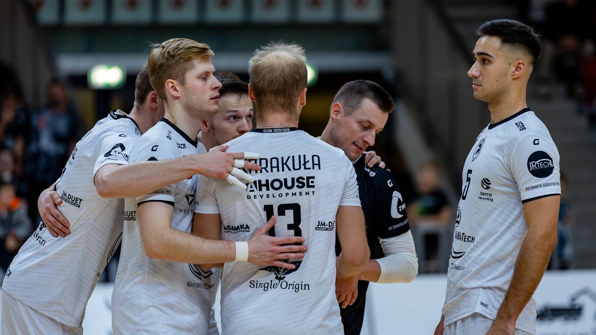 Grupa siatkarzy w białych koszulkach przytulająca się na boisku.