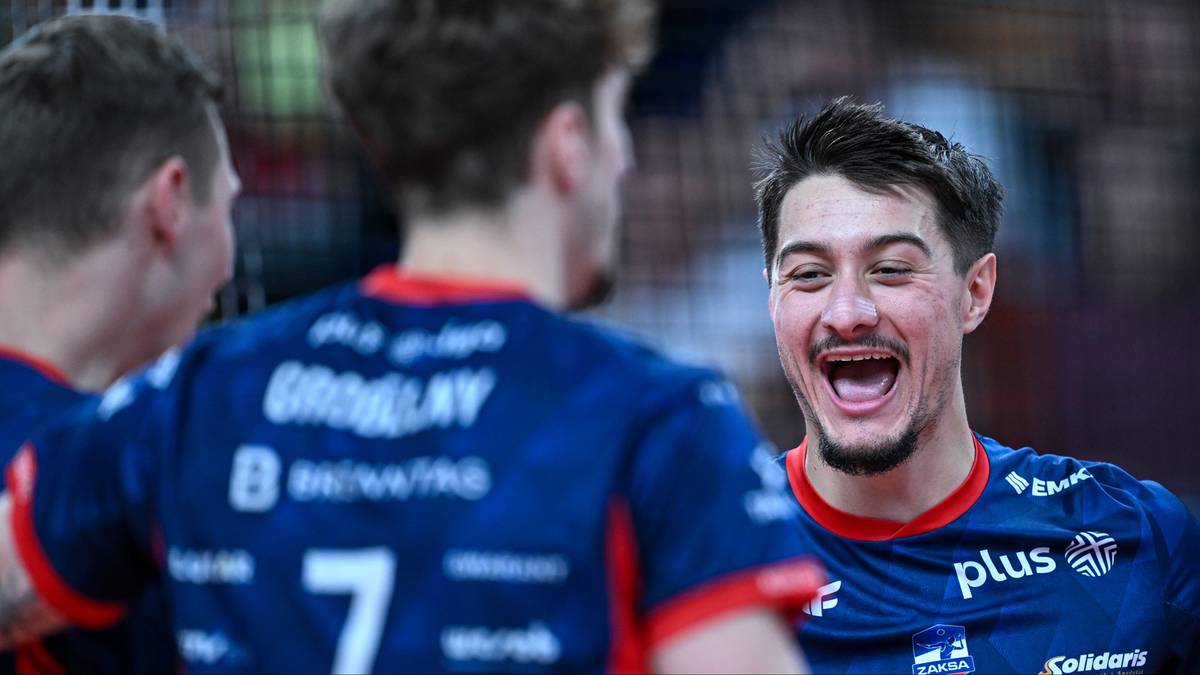 Trzech siatkarzy w niebiesko-czerwonych strojach sportowych, jeden z nich szeroko uśmiechnięty, celebruje zwycięstwo na boisku.