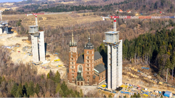 Dwie wysokie, nowoczesne konstrukcje wsporcze estakady po obu stronach bazyliki Mariackiej w Krakowie.