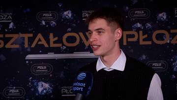 Młody mężczyzna w czarnej kamizelce i białej koszuli z kołnierzykiem m&oacute;wi do mikrofonu z logo Polsat Sport.