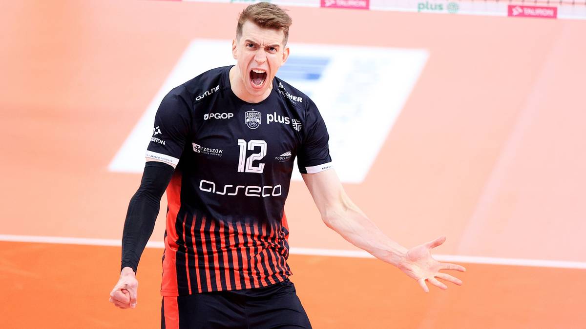 Siatkarz w czarno-pomarańczowej koszulce z numerem 12, krzyczy z radości na boisku siatkarskim.