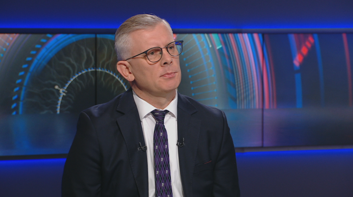 Mężczyzna w okularach i garniturze siedzi i patrzy w bok.