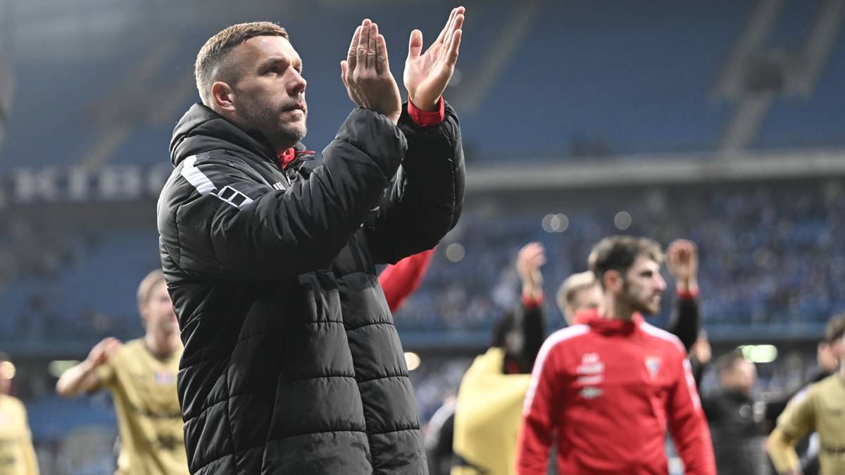 Mężczyzna w sportowej kurtce klaszcze w dłonie na tle innych os&oacute;b i trybun stadionu.