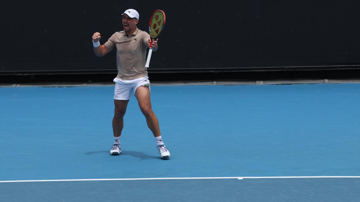 Tenisista w białej czapce, beżowej koszulce i białych spodenkach z rakietą w ręku, triumfalnie unosi pięść na korcie tenisowym.