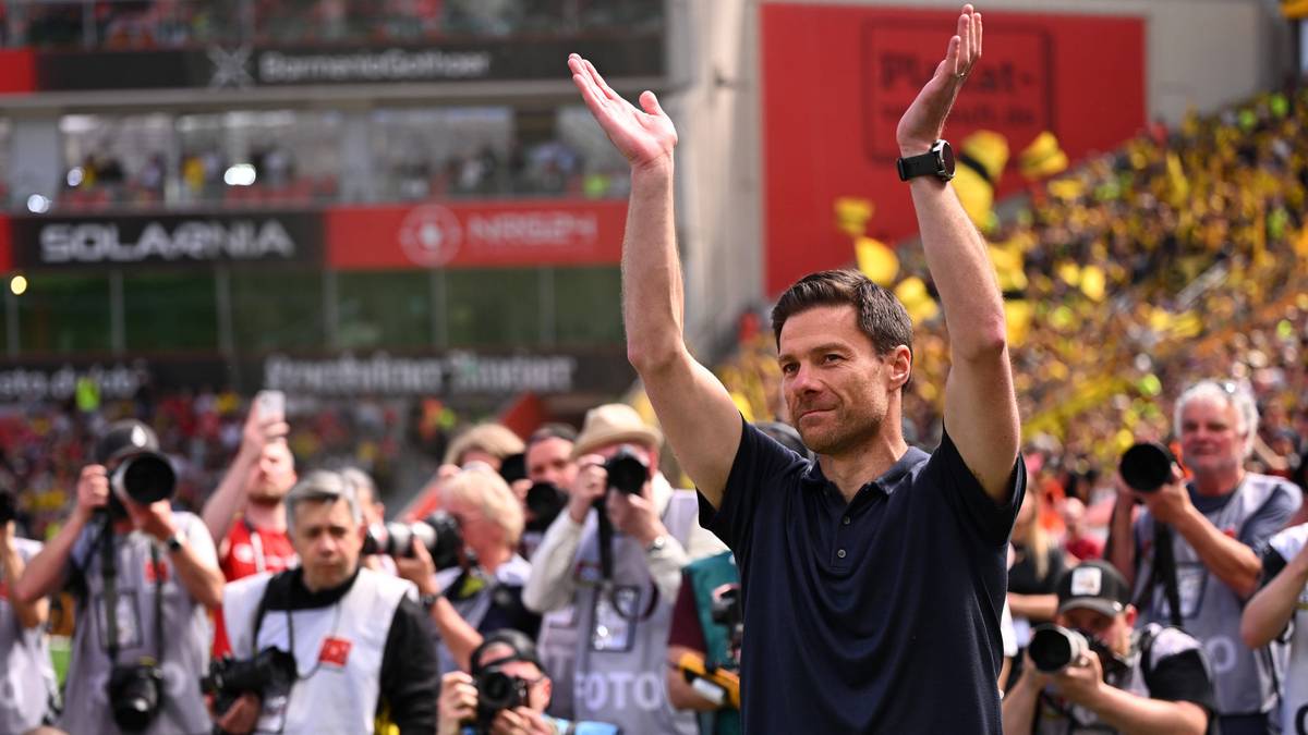 Uśmiechnięty mężczyzna w czarnej koszulce, Xabi Alonso, podnosi ręce w geście pozdrowienia do tłumu. W tle widać innych ludzi, reporterów z aparatami i trybuny stadionu.