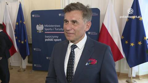 Robert Korzeniowski w garniturze, stojący przed panelem Ministerstwa Sportu i Turystyki, z polskimi i unijnymi flagami w tle.