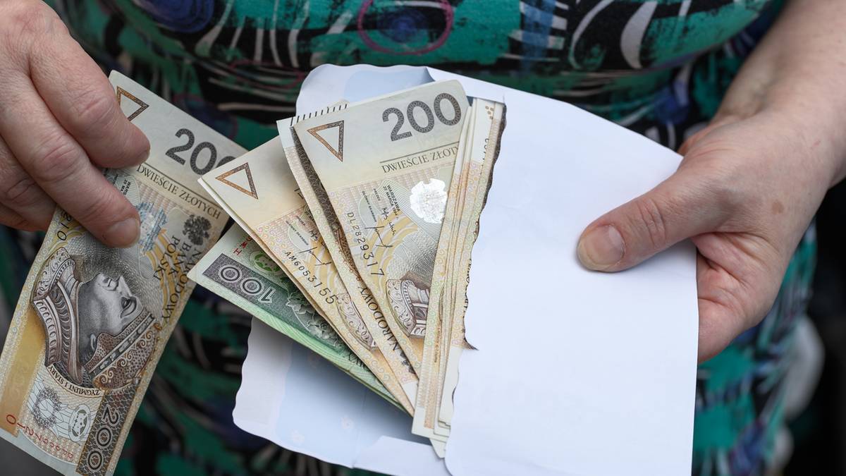 Dłonie trzymające kopertę z polskimi banknotami 200 zł i 20 zł.