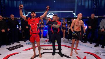 Dwóch zawodników MMA stoi na macie, jeden w czerwonym stroju z uniesionymi rękami świętuje zwycięstwo, drugi w czarnym stroju stoi obok.