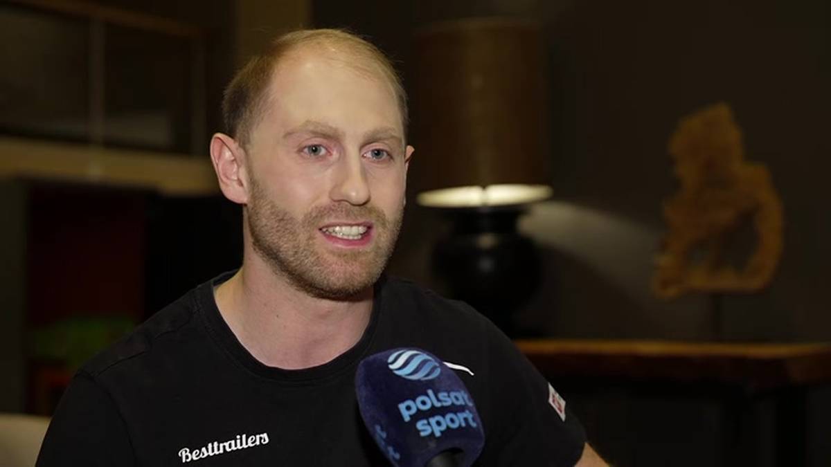 Mężczyzna z zarostem i łysiejącą głową, ubrany w czarną koszulkę, m&oacute;wi do mikrofonu z logo Polsat Sport. W tle widać lampę i ozdobne przedmioty.