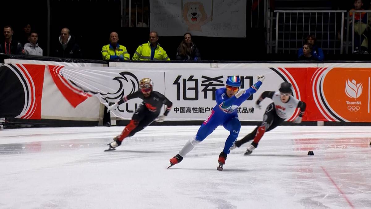 Trzech łyżwiarzy szybkich rywalizuje na lodzie. Zawodnik w niebieskim stroju prowadzi, a za nim jadą zawodnicy w czerwonym i czarnym stroju.