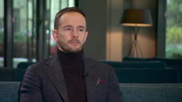 Mężczyzna w ciemnym golfie i marynarce siedzi w fotelu.