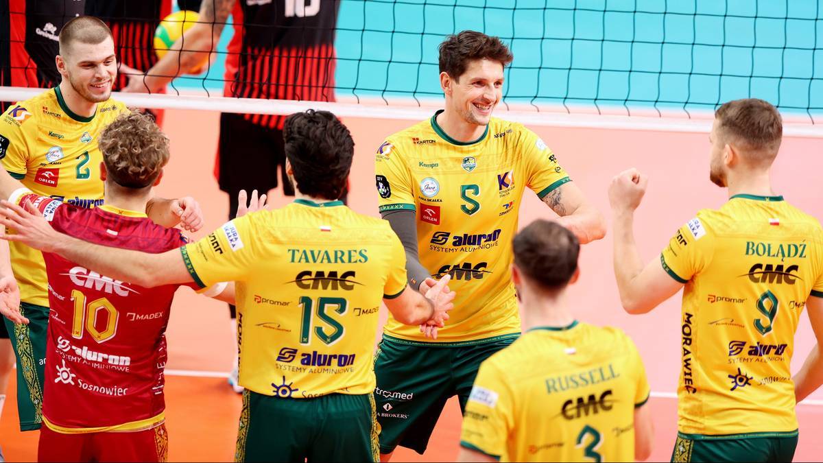 Grupa siatkarzy w ż&oacute;łtych i czerwonych strojach sportowych raduje się po udanej akcji na boisku siatkarskim.