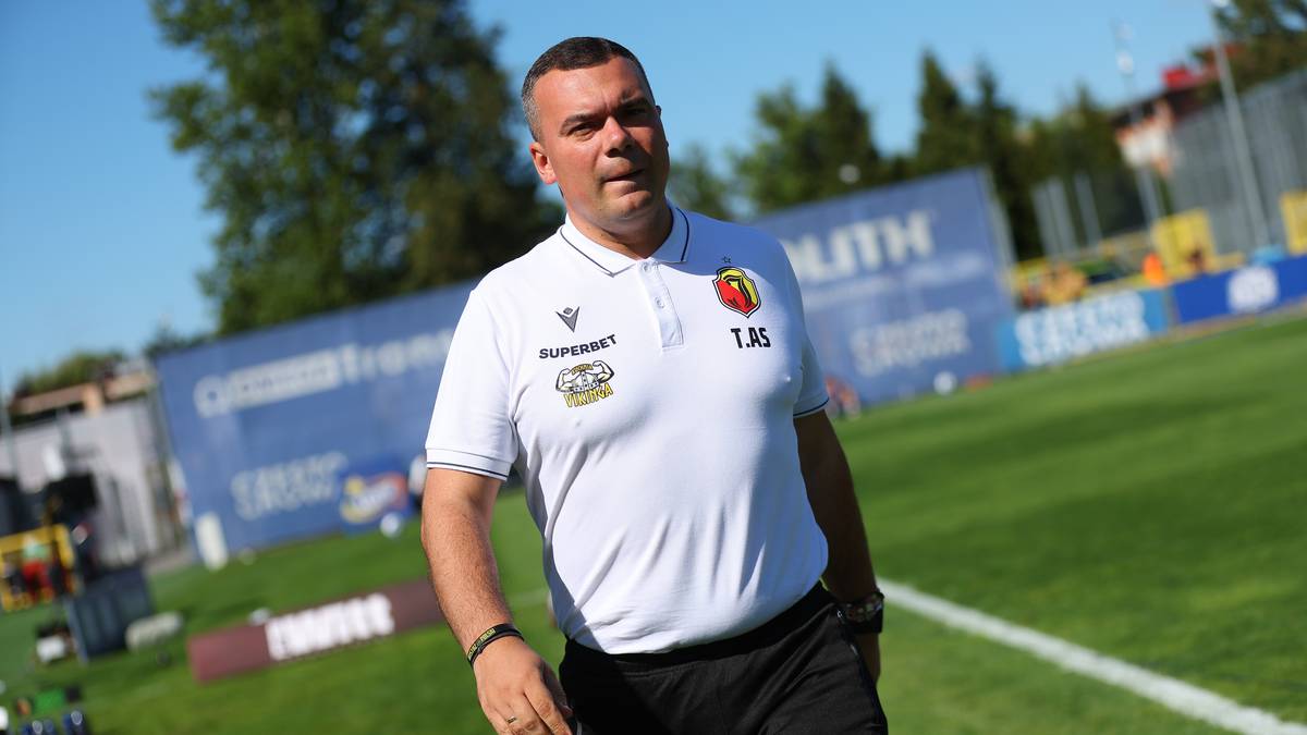 Mężczyzna w białej koszulce polo z herbem klubu sportowego, w okularach przeciwsłonecznych, idzie po boisku piłkarskim.