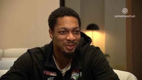 Mężczyzna o ciemnej karnacji i z kr&oacute;tkimi, kręconymi włosami, ubrany w czarną bluzę z kapturem, uśmiecha się do kamery. W tle widoczna lampa i jasne ściany.