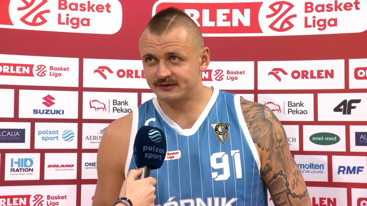 Koszykarz z numerem 91 na koszulce, z charakterystyczną fryzurą i wąsami, udziela wywiadu na tle ścianki sponsorskiej.