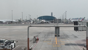 Widok na płytę lotniska w Dubaju z samolotami pasażerskimi i budynkiem terminala w tle.