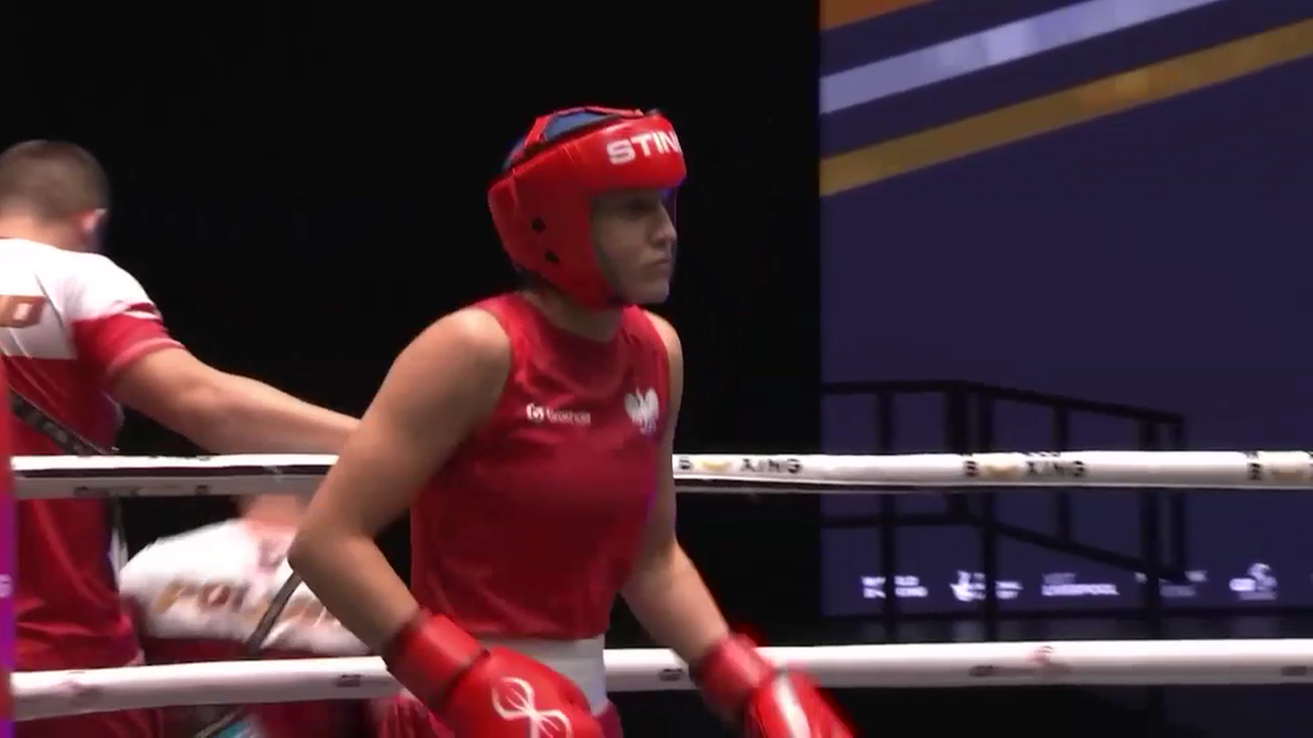 Bokserka w czerwonym stroju i kasku zbliża się do narożnika ringu, podczas gdy druga osoba w białej koszulce z biało-czerwonym pasem jest częściowo widoczna obok niej.