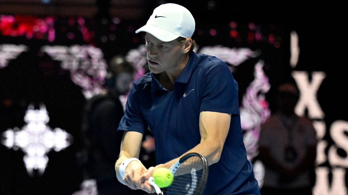 Tenisista Jannik Sinner w białej czapce i granatowej koszulce przygotowuje się do serwisu, trzymając rakietę i piłkę.