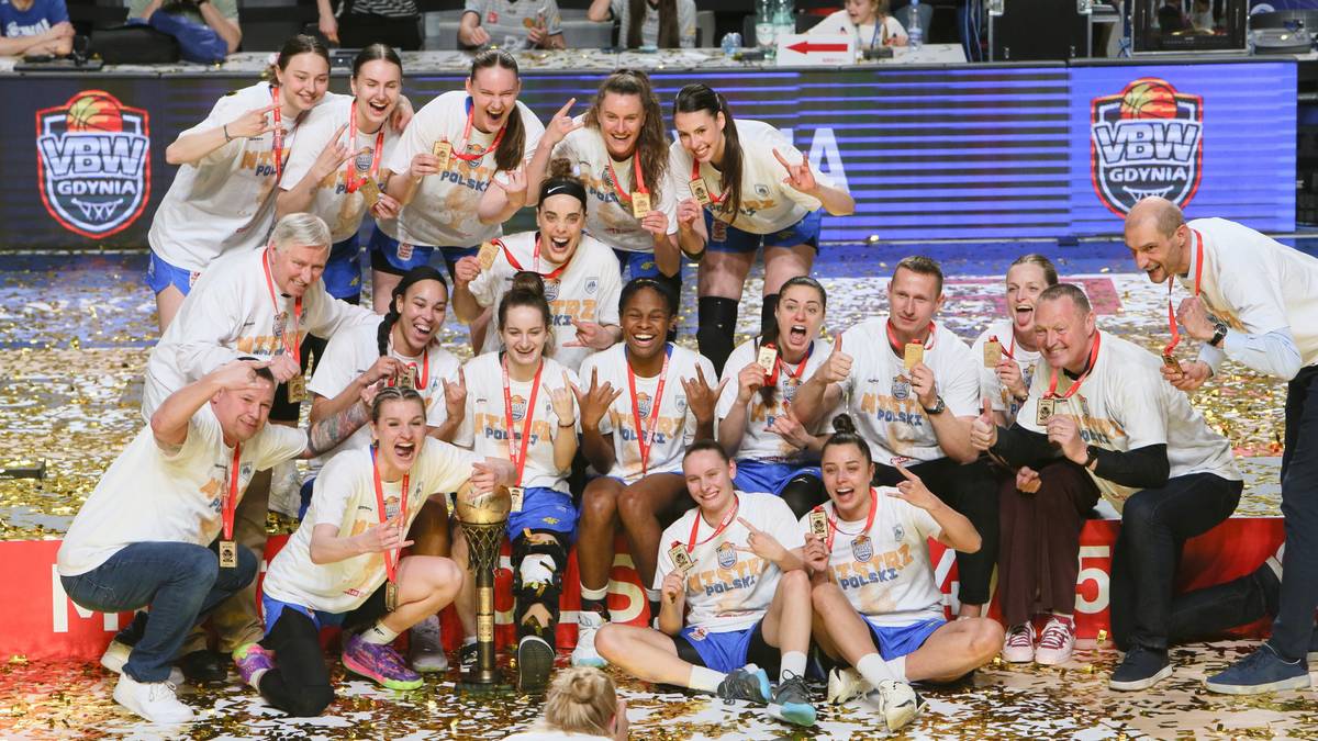 Drużyna koszykarek w białych koszulkach i spodenkach, trzymająca złote trofeum i otoczona konfetti, celebrująca zwycięstwo.